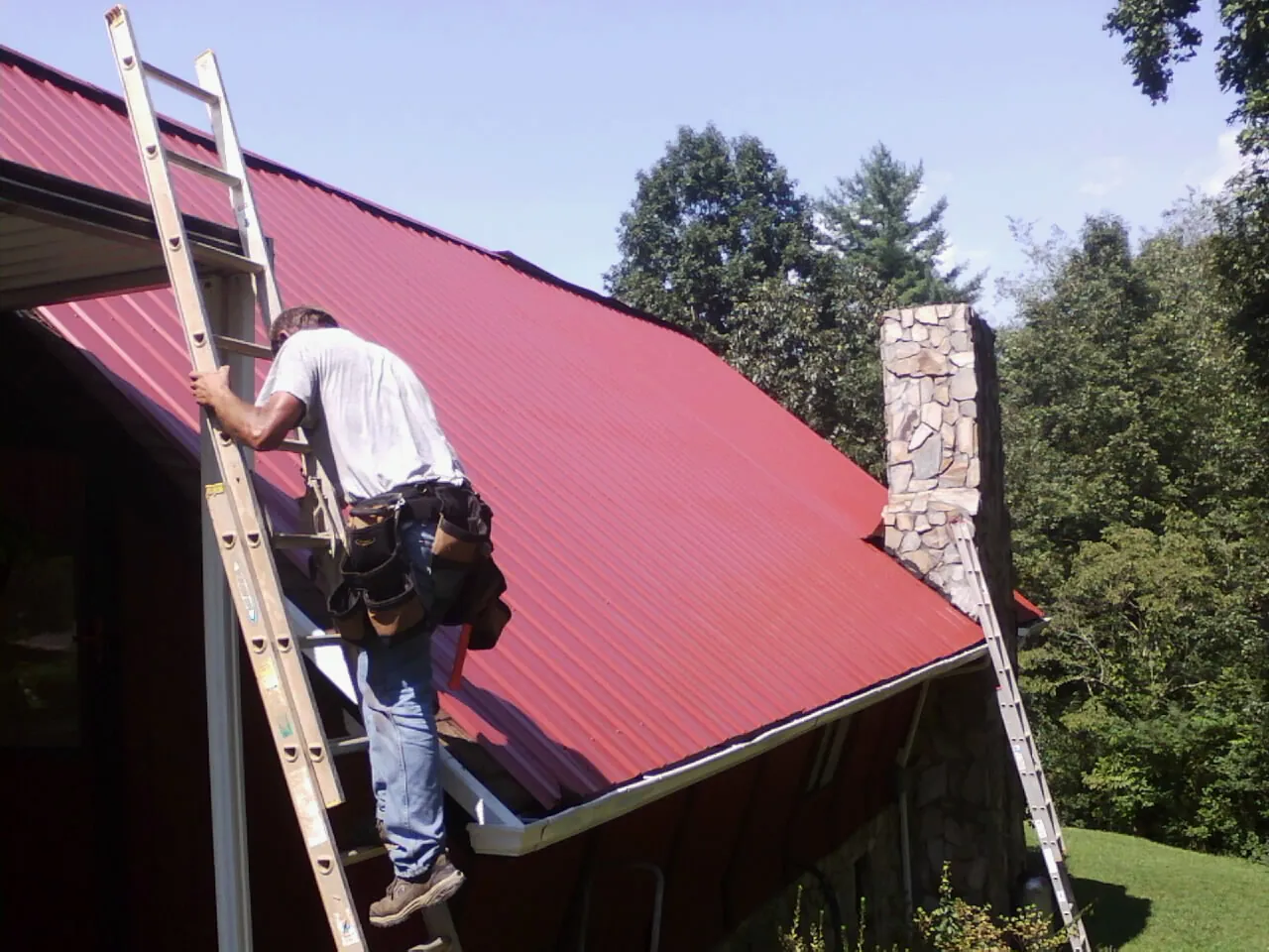 Roofer on a ladder beside a red metal roof and stone chimney — Soffit & Fascia Repair by Roofing Brothers in Waitsburg